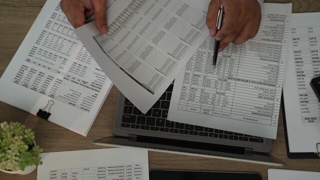 Accountant reviewing printed financial reports with pen over laptop for accounting, tax preparation, audit, bookkeeping, financial planning during tax season.