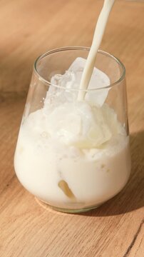Oat milk pours over ice cubes in clear glass during preparation of refreshing iced hojicha latte on warm wooden tabletop.