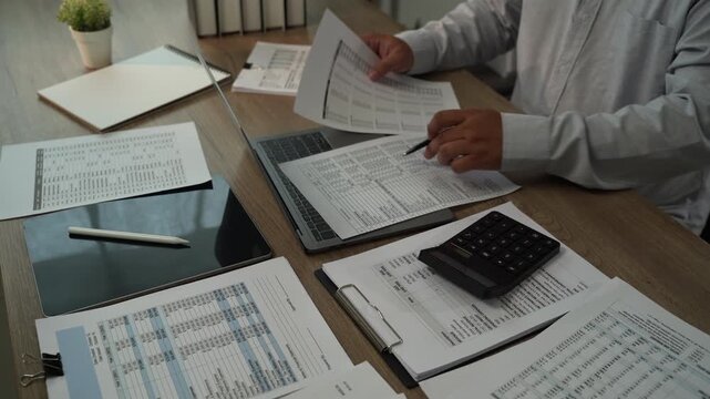 Accountant using calculator while reviewing financial reports at office desk for accounting, tax preparation, audit, bookkeeping, financial planning.