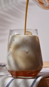 Hojicha tea pours into oat milk and ice in clear glass, creating layered iced latte with golden syrup on wooden tabletop.