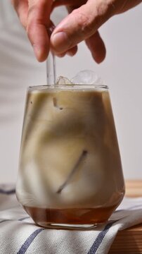 Hand stirs iced hojicha latte with oat milk and syrup in clear glass during preparation of a creamy chilled drink at home.