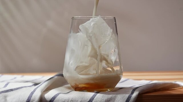 Oat milk pours over ice and golden syrup in clear glass during preparation of a chilled homemade drink on wooden tabletop.