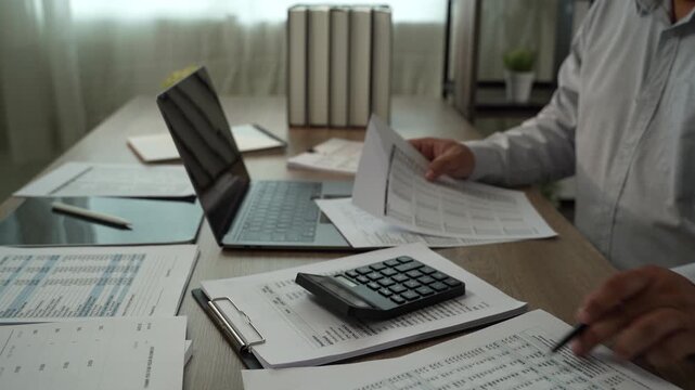 Accountant using calculator while reviewing financial reports with laptop, tablet, pen for accounting, tax preparation, audit, bookkeeping, financial planning.