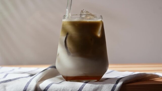 Hand stirs hojicha with oat milk and ice in clear glass, blending layered iced latte on wooden table in close-up at home.