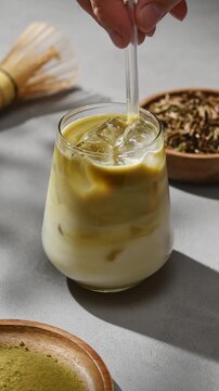 Hand stirs iced hojicha latte with glass straw as roasted tea blends into oat milk beside bamboo whisk and tea powder gently.