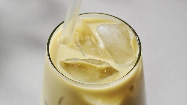 Hand stirs iced hojicha latte with oat milk in clear glass, mixing creamy roasted tea drink with ice on light background.