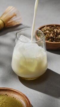 Oat milk pours over ice in glass beside hojicha powder, tea leaves and bamboo whisk during preparation of chilled tea latte.