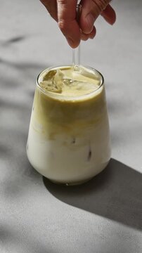Hand stirs iced hojicha latte with glass straw, blending roasted tea and oat milk into creamy cold beverage in soft sunlight.