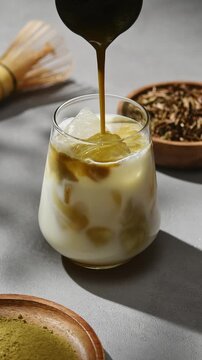 Hojicha tea pours over oat milk and ice, forming layered iced latte beside bamboo whisk, roasted leaves and green tea powder.