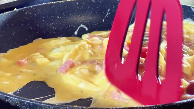 Close-up shot of a red spatula stirring an omelette with diced ham and tomatoes in a skillet. Ideal for breakfast recipes, cookware ads, and culinary videos.