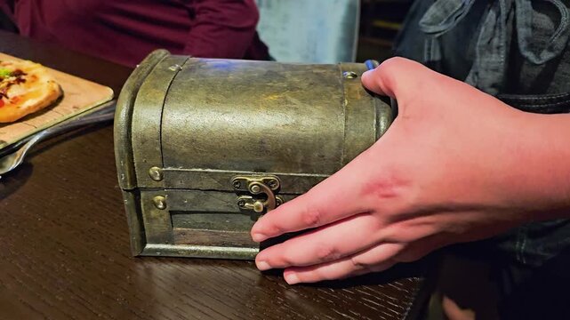 Hand closes a rustic wooden presentation box over a smoking cocktail and blue popsicle garnish on a restaurant table, with dark wood, moody bar lighting, and dramatic dining atmosphere.