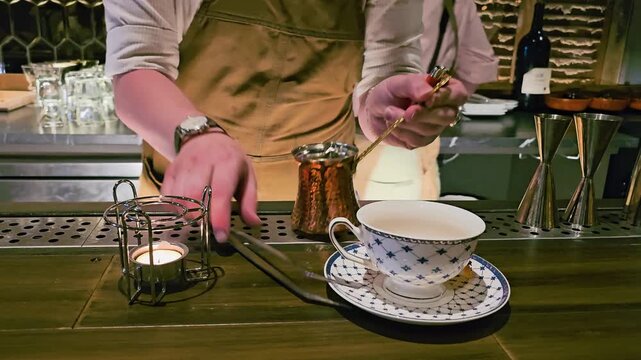 Close view of copper pot being heated for specialty tea service, with candle flame, decorative teacup, saucer, tweezers, bar tools, and soft evening lounge atmosphere.