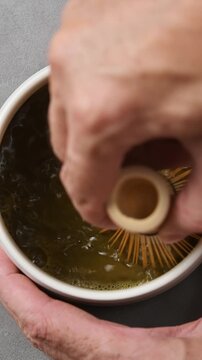 Hands whisk roasted hojicha tea with bamboo chasen in ceramic bowl, creating fine foam during traditional drink preparation.