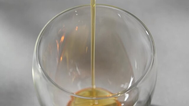 Golden syrup pours into clear double wall glass before hojicha latte preparation in horizontal closeup on neutral background.