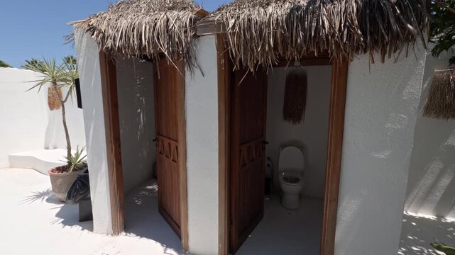 Outdoor restroom stalls with red door frames and a thatched palm roof stand at a sunny white washed beach resort
