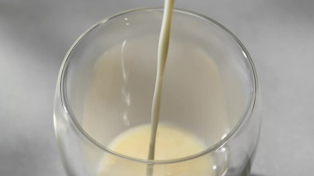 Milk pours over syrup into clear glass during hojicha latte preparation in horizontal closeup on neutral kitchen background.