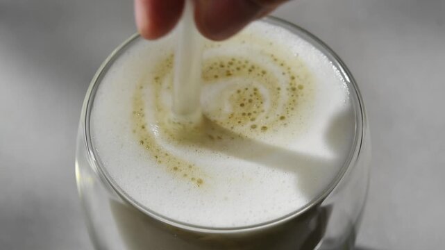 Hojicha concentrate is stirred into foamed milk in clear glass, blending roasted tea latte in horizontal macro closeup view.