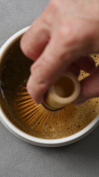 Bamboo whisk moves slowly across foamy hojicha surface in ceramic bowl, showing rich tea texture in vertical macro closeup.