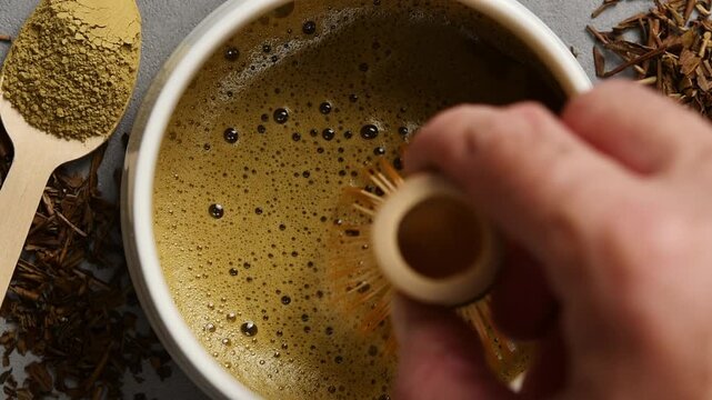 Hand slowly moves bamboo whisk across foamy hojicha in ceramic bowl to show creamy texture and fine bubbles in close view.