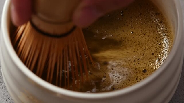 Bamboo whisk rapidly froths hojicha in ceramic bowl, showing rich foam texture and swirling surface in close up macro view.