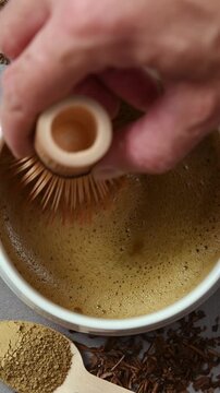 Hand slowly moves bamboo whisk through foamy hojicha in ceramic bowl, revealing smooth texture during traditional tea making.