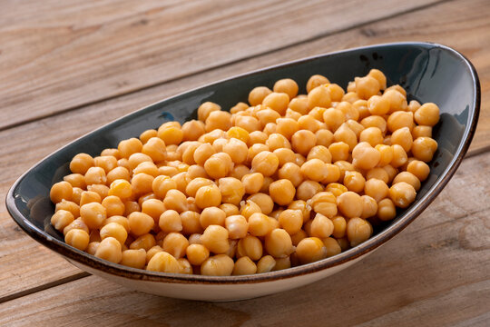 Boiled chickpeas heap on an oval ceramic platter. Fresh organic garbanzo beans, close-up on a rustic dark wood plank background, in a tilted view with natural window light.