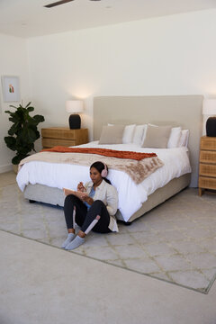 African American woman wearing leggings sitting in bedroom using tablet and stylus with headphones