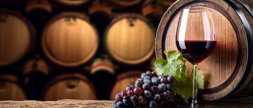 Red wine glass and grapes on oak barrel with blurred cellar background banner.