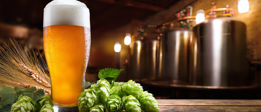 Glass of amber craft beer with hop cones and blurred brewery background banner.