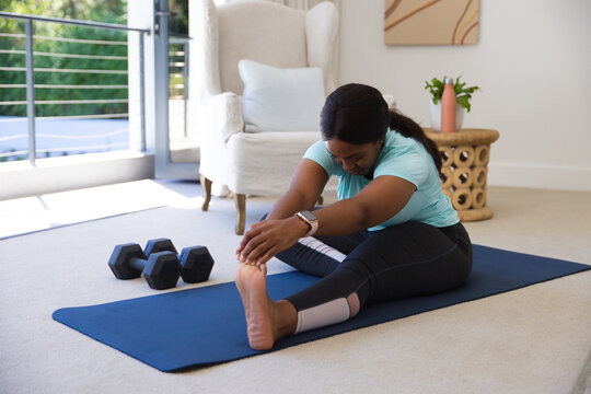 African American woman doing hamstring stretch on blue mat at home reaching toes dumbbells near