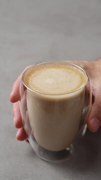 Hand gently places glass of creamy hojicha oat milk latte on gray table, showing finished Japanese tea drink ready to serve.