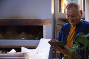 Fototapeta premium Senior Asian man holding tablet and standing in living room near sofa by fireplace, copy space