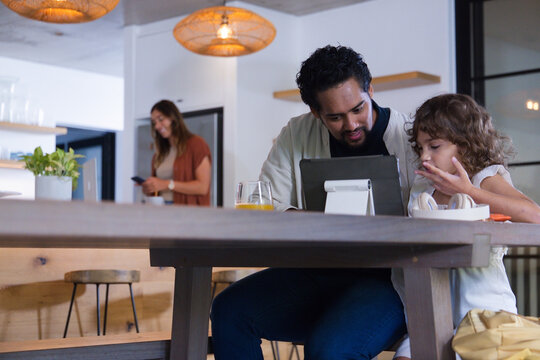 African American man and child leaning toward tablet at wooden table with headphones eating snack