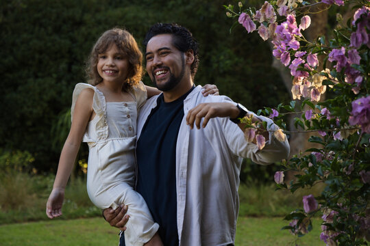 African American father holding daughter in garden, smiling, wearing wristwatch, near blooms