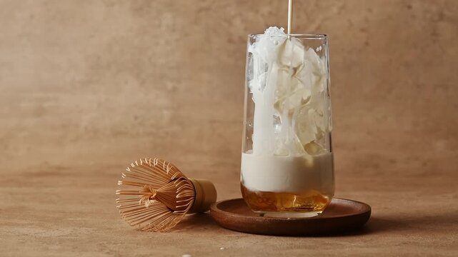 Oat milk pours over ice and syrup in tall glass beside bamboo whisk, forming creamy base for cold hojicha latte preparation.