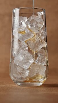 Golden syrup pours over ice cubes in tall glass, coating chilled surface during preparation of sweet iced beverage close up.