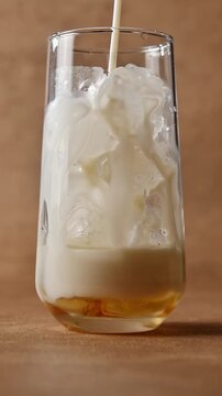 Cold oat milk pours over ice and syrup in tall glass, creating layered base for creamy iced beverage preparation close up.