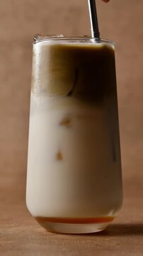 Hand stirs layered hojicha latte with straw, mixing roasted tea, oat milk and syrup into creamy iced drink in tall glass.
