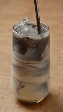 Hand stirs black sesame paste into iced oat milk, blending dark layers into creamy black sesame latte inside clear glass.