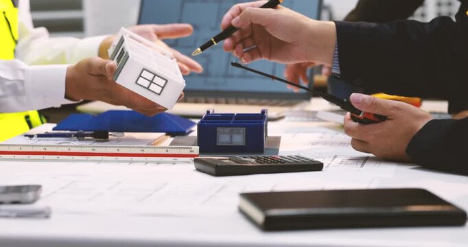 Architects and engineers review a small house model, blueprints, ruler and calculator during a building design meeting at a desk.