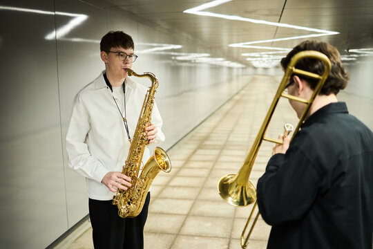 Two young adult Caucasian male street musicians playing saxophone and trombone in modern pedestrian underpass, representing urban culture and creative performance for music promotion