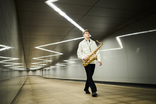 Young adult Caucasian man walking through modern pedestrian underpass holding saxophone, representing street musicians. Image suitable for music education and urban lifestyle marketing