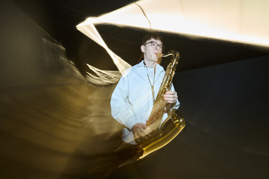 Young adult Caucasian man playing saxophone on street at night with motion blur light trails. Image suitable for jazz festival promotion or urban music culture marketing, low angle shot