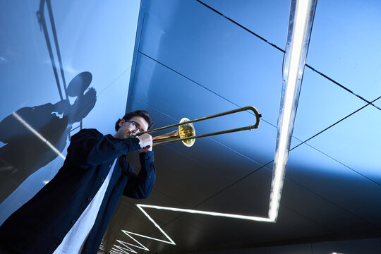 Young adult Caucasian man playing trombone in modern underpass, casting shadow on wall. Image suitable for music education promotion and urban lifestyle marketing, low angle shot
