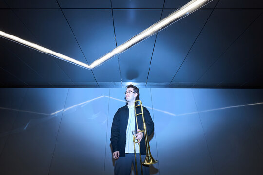 Young adult Caucasian male street musician holding trombone in modern underpass with neon lighting, representing urban art culture. Ideal for music event promotion, low angle shot