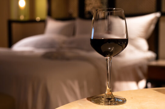 Glass of red wine on a table with a bedroom bed in the background.