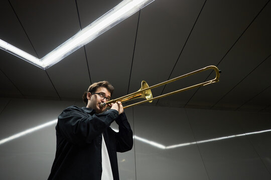 Young adult Caucasian man playing trombone in underground passage, representing street musicians and urban culture. Image suitable for music education and arts promotion, low angle shot