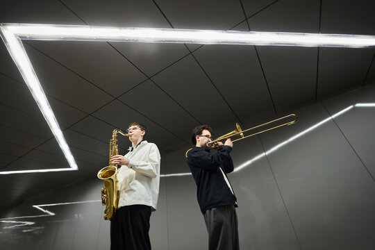Two young adult Caucasian male street musicians playing saxophone and trombone in modern pedestrian underpass. Image suitable for music education and urban lifestyle marketing, low angle view