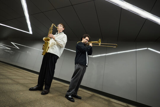 Two young adult Caucasian male street musicians playing saxophone and trombone in modern underground passage. Image suitable for urban culture and live music promotion, wide angle shot