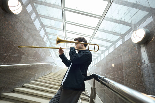 Young adult Caucasian man playing trombone on stairs in underpass, performing as street musician. Image suitable for urban culture articles and music education promotions, wide angle shot